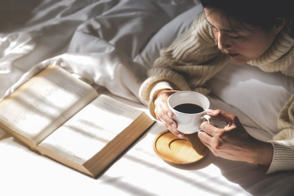 Asian woman holding cup of coffee and reading a book on the bed after get up in morning