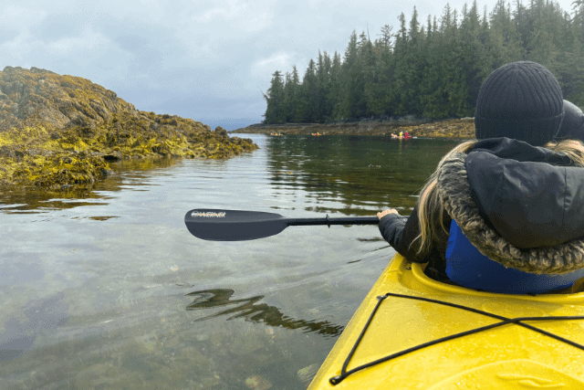 Person paddling a yellow kayak in a calm coastal waterway, surrounded by rocky outcrops and evergreen trees on a cloudy day.