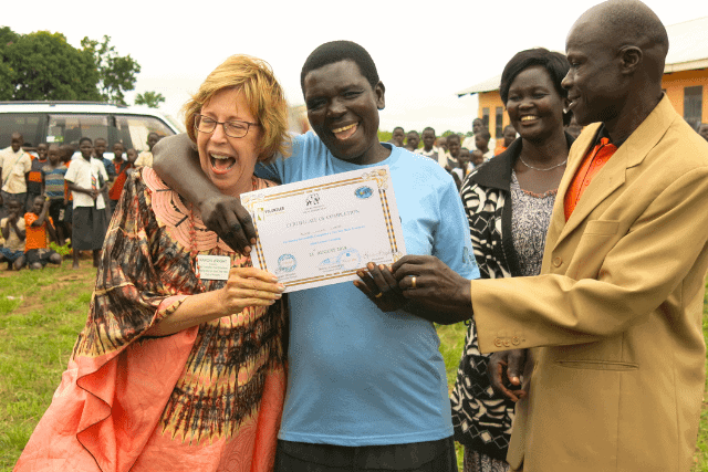 A group of people celebrate as one person holds a certificate. Another person laughs with an arm around them. There are onlookers and a vehicle in the background.