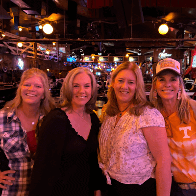 Four women standing together in a dimly lit venue with wooden decor and string lights.