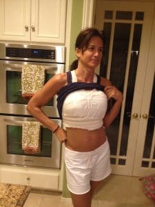 A woman in a kitchen lifts her shirt to show her chest wrapped in thick white bandages, possibly for medical reasons. She is wearing white shorts and a navy-blue tank top.