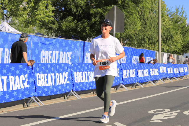 A woman running in a race with a "Great Race" banner in the background, smiling.
