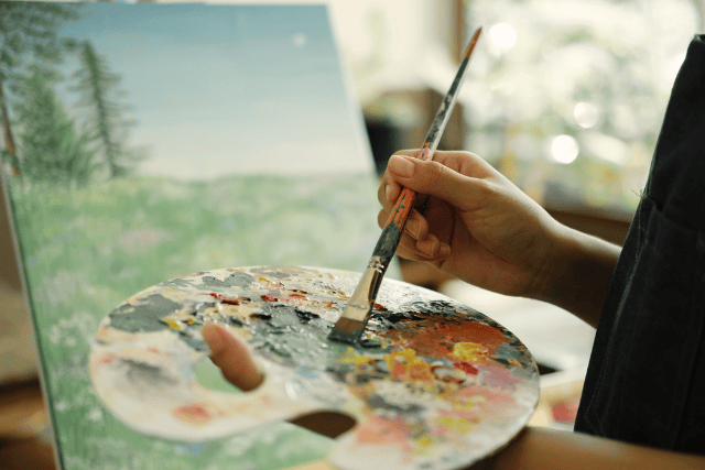 Artist's hand holding a paintbrush and palette with colorful paints in a bright studio.